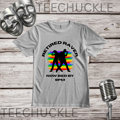 A gray graphic t-shirt displayed on a rustic wooden background, featuring a black silhouette of a couple on a rainbow sunset with the text 'RETIRED RAVER NOW BED BY 9PM'. The image includes decorative props like a vintage microphone, comedy and tragedy masks, and a silver microphone stand.