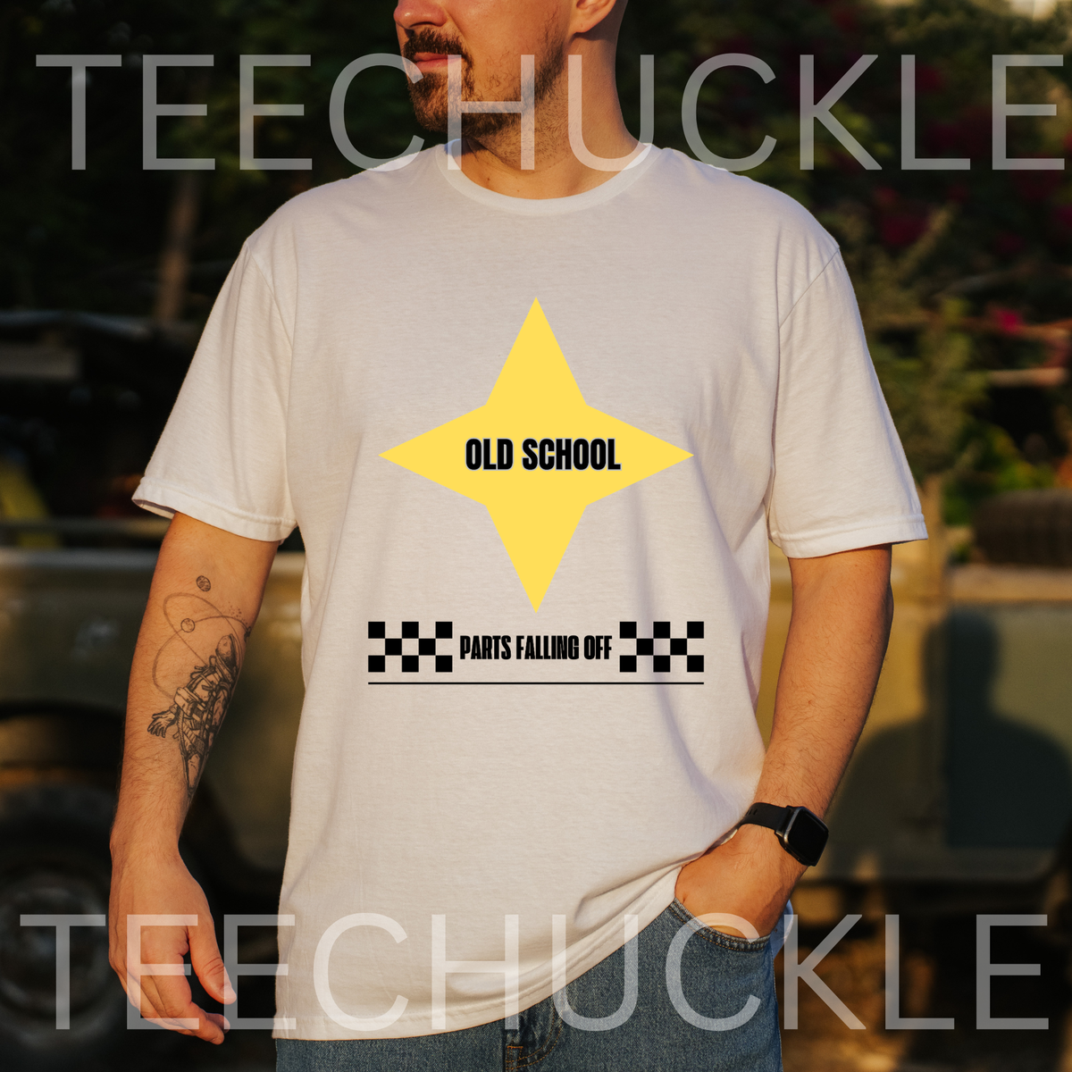 A man wearing a white graphic t-shirt with a yellow star and black text reading 'OLD SCHOOL' and 'PANTS FALLING OFF'. The background is a blurred outdoor setting with a vehicle
