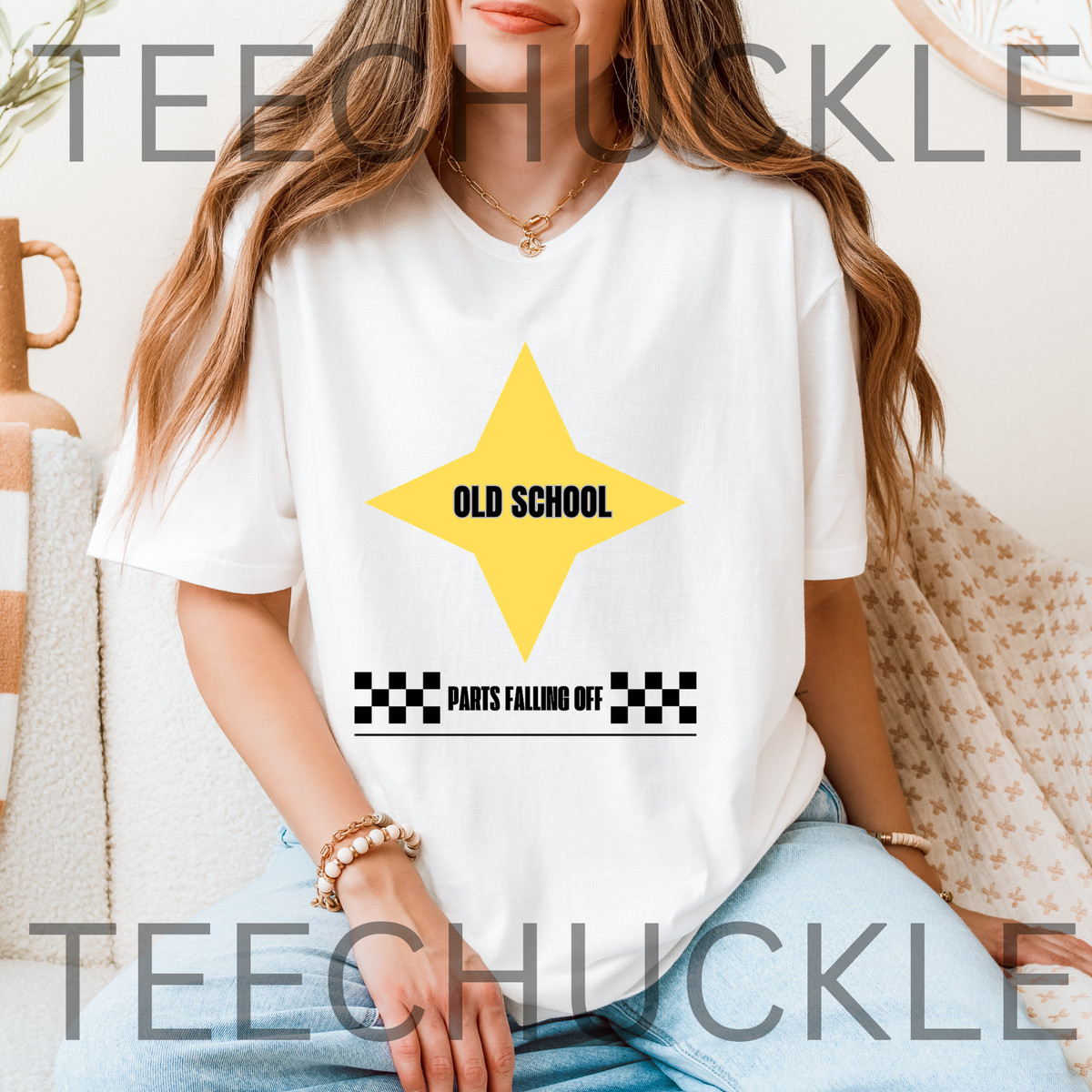 A woman wearing a white graphic T-shirt with a yellow star and the text 'OLD SCHOOL' and 'PARTS FALLING OFF' on a checkered strip, seated indoors.