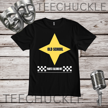 A black short-sleeve graphic T-shirt laid flat on a rustic wood plank background, with a large yellow star graphic and the words 'OLD SCHOOL' and 'PARTS FALLING OFF' printed on the front. Silver theater masks and a vintage microphone are placed around the shirt as styled props.