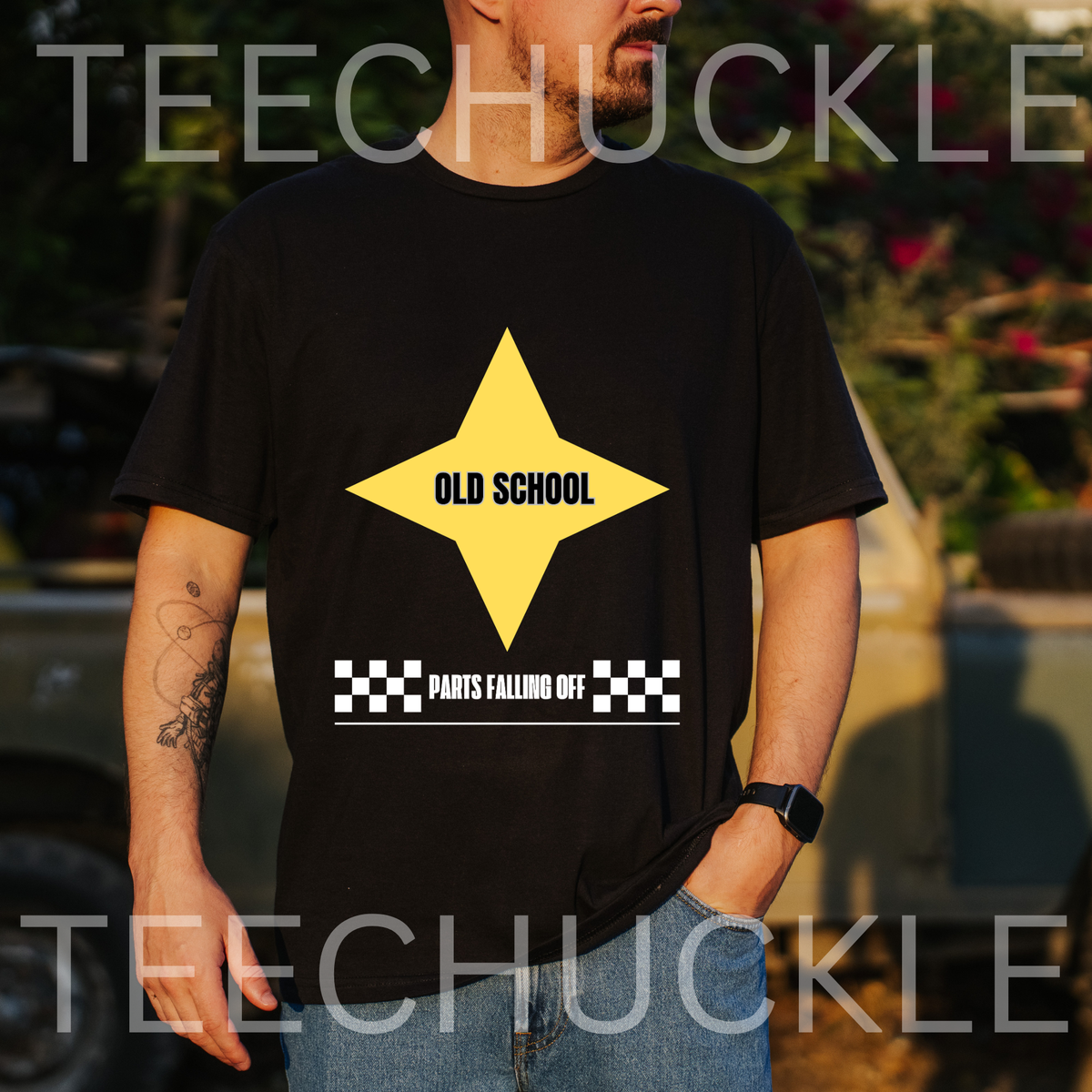 A man wearing a black graphic t-shirt with a yellow star and the text 'OLD SCHOOL' and 'PARTS FALLING OFF'. The background is an outdoor setting with a blurred truck and foliage.