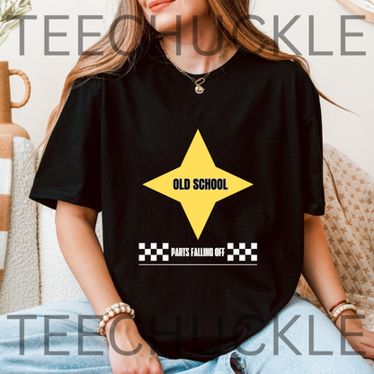A woman wearing a black oversized T-shirt with a yellow star graphic and the words 'OLD SCHOOL' and 'PARTS FALLING OFF' printed on the front. The photo is taken in a cozy indoor setting with a textured beige couch and a ceramic vase in the background.