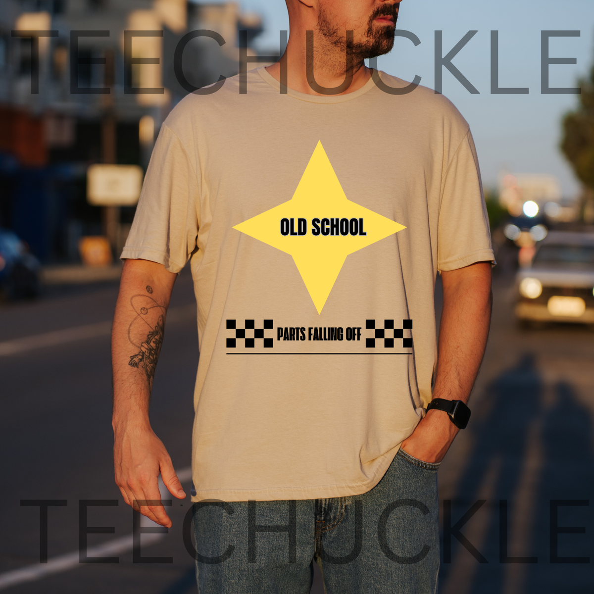 A man wearing a beige graphic t-shirt with a yellow star and black text reading 'OLD SCHOOL' and 'PARTS FALLING OFF', photographed outdoors on a blurred street background.