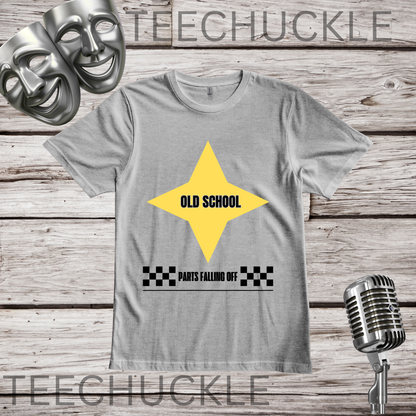 A gray crewneck t-shirt with a yellow star graphic containing the text 'OLD SCHOOL' and a black-and-white checkered bar below reading 'PARTS FALLING OFF'. The shirt is displayed on a rustic wooden background with a vintage microphone, comedy masks.