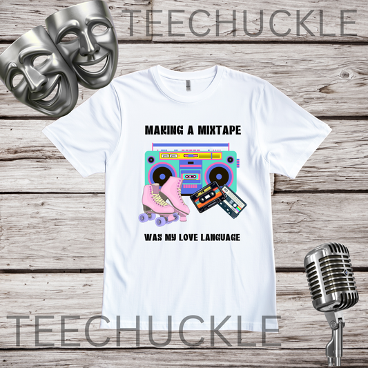 A white graphic t-shirt displayed on a rustic wood background, featuring a retro boombox, cassette tape, and pink roller skate design with the text 'MAKING A MIXTAPE WAS MY LOVE LANGUAGE'. Two silver comedy masks and a vintage microphone are styled as props.
