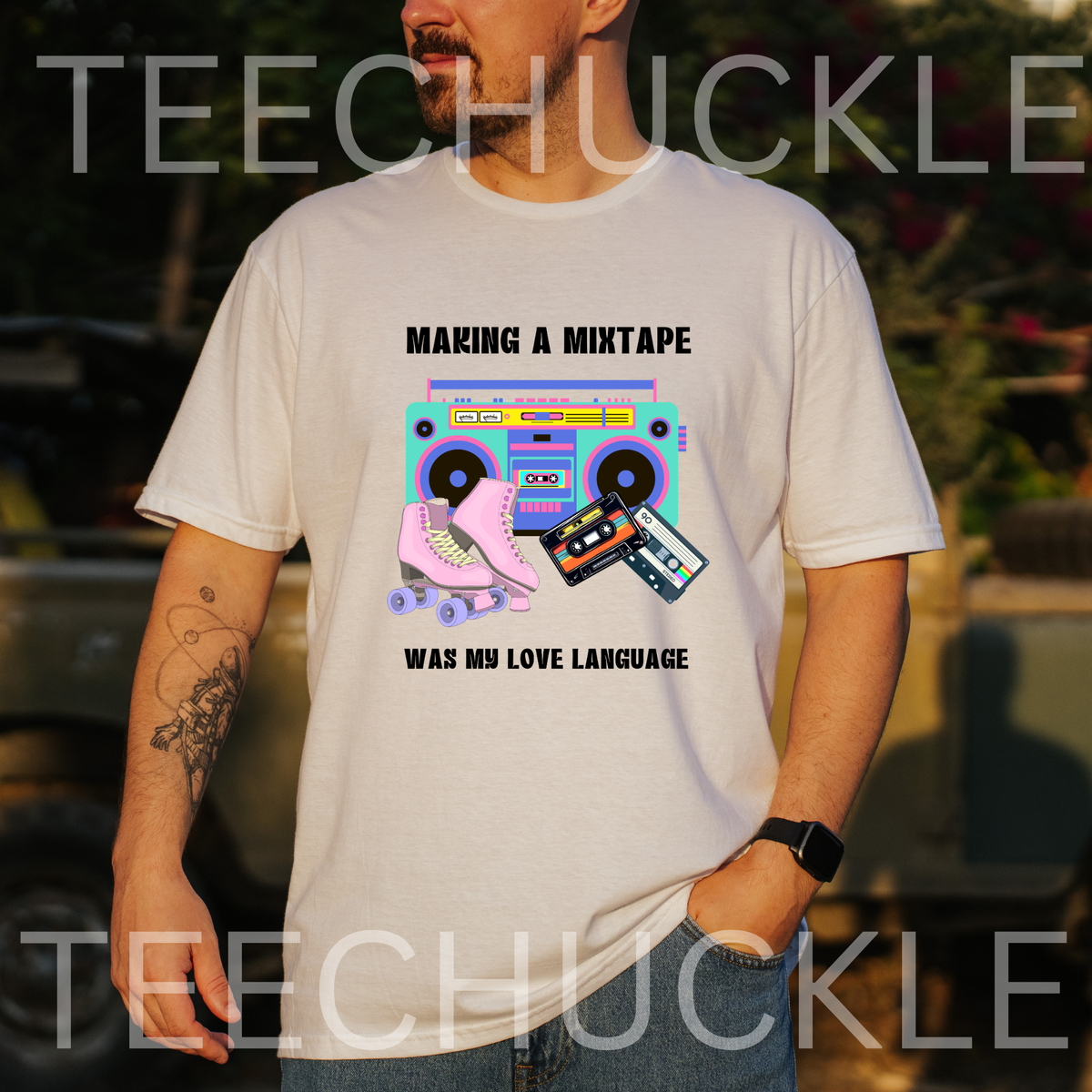 A man wearing a white graphic t-shirt with a retro cassette tape and boombox design that reads 'MAKING A MIXTAPE WAS MY LOVE LANGUAGE'. The background is a blurred outdoor setting with warm sunset lighting