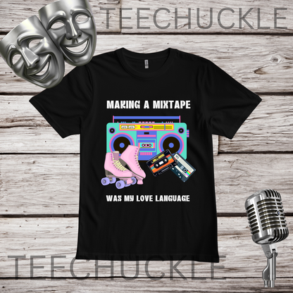 A black short-sleeve graphic T-shirt laid flat on a rustic wood background, with two silver theater masks, a vintage microphone. The shirt graphic reads 'MAKING A MIXTAPE WAS MY LOVE LANGUAGE' and shows a retro boombox, cassette tapes, and a pink roller skate.