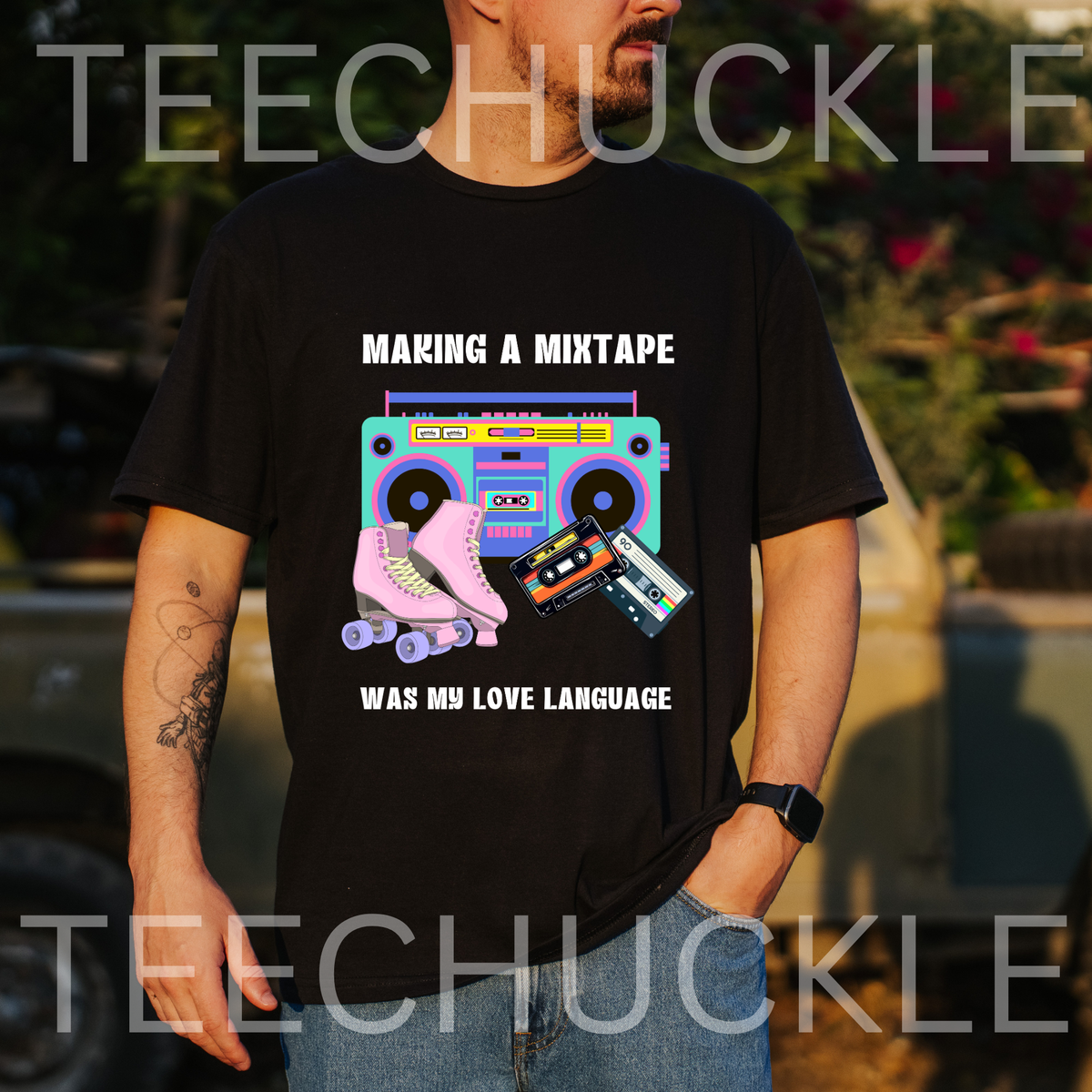 A man wearing a black graphic t-shirt with a retro 80s/90s boombox, cassette tape, and roller skate design, with text "making a mixrape was my love language" standing outdoors in front of a blurred vehicle.