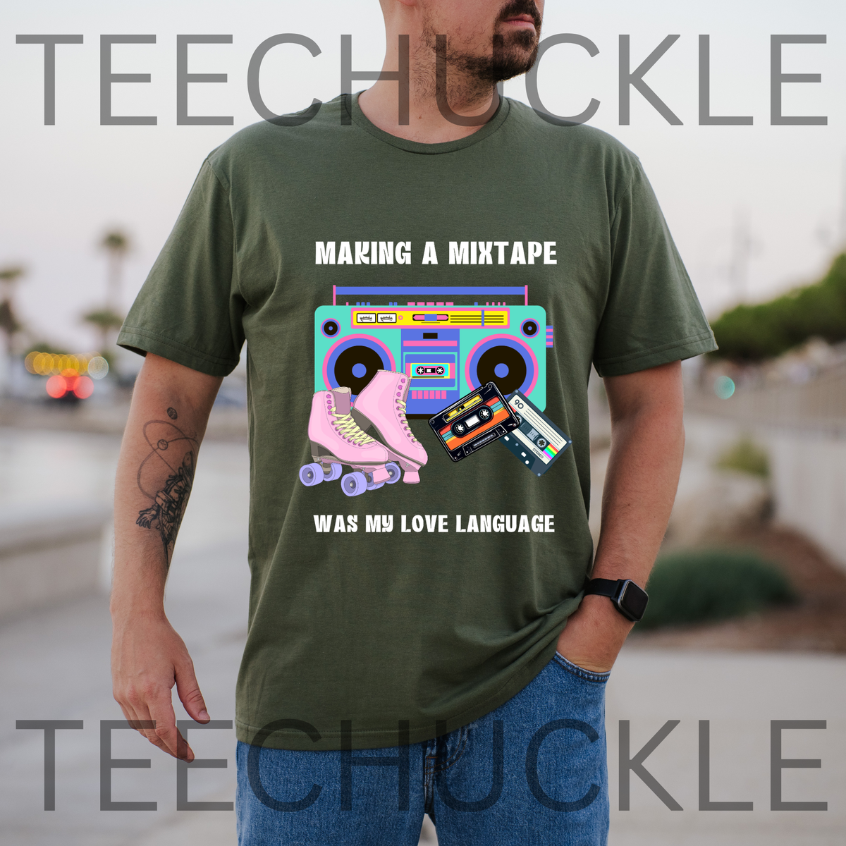 A man wearing a green graphic t-shirt with a retro cassette tape and roller skate design, standing outdoors with a blurred background. The t-shirt features the text 'MAKING A MIXTAPE WAS MY LOVE LANGUAGE' and a colorful retro boombox and cassette graphic.