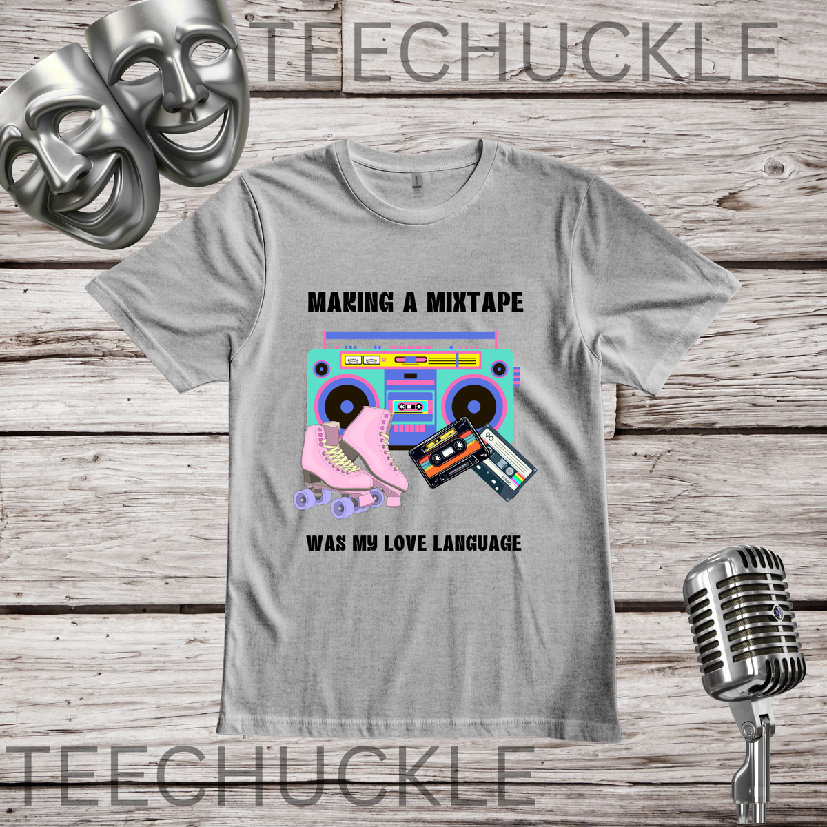 A gray graphic t-shirt displayed on a rustic wooden background, featuring a retro 80s design with a pink roller skate, cassette tapes, and a boombox, with the text 'MAKING A MIXTAPE WAS MY LOVE LANGUAGE', with a silver microphone and comedy masks.