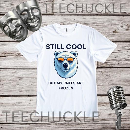 A white graphic t-shirt displayed on a rustic wood background, featuring a polar bear illustration wearing orange-tinted sunglasses with the text 'STILL COOL BUT MY KNEES ARE FROZEN', and styled with silver comedy and tragedy masks and a vintage microphone.