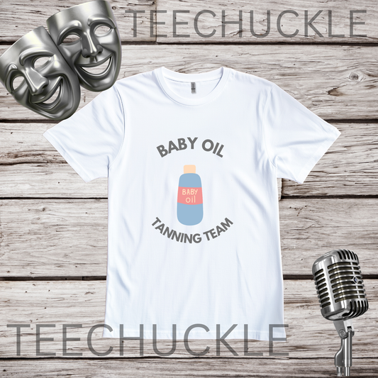 A white graphic t-shirt featuring a 'Baby Oil Tanning Team' design with a bottle illustration, displayed on a rustic wood background with silver theater masks and a vintage microphone. 