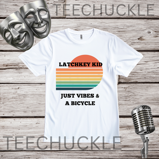 A white graphic t-shirt displayed on a rustic wood background, featuring a retro sunset design with the text 'LATCHKEY KID JUST VIBES & A BICYCLE'. The shirt is styled with two silver theatrical masks in the top left and a vintage microphone on the bottom right.