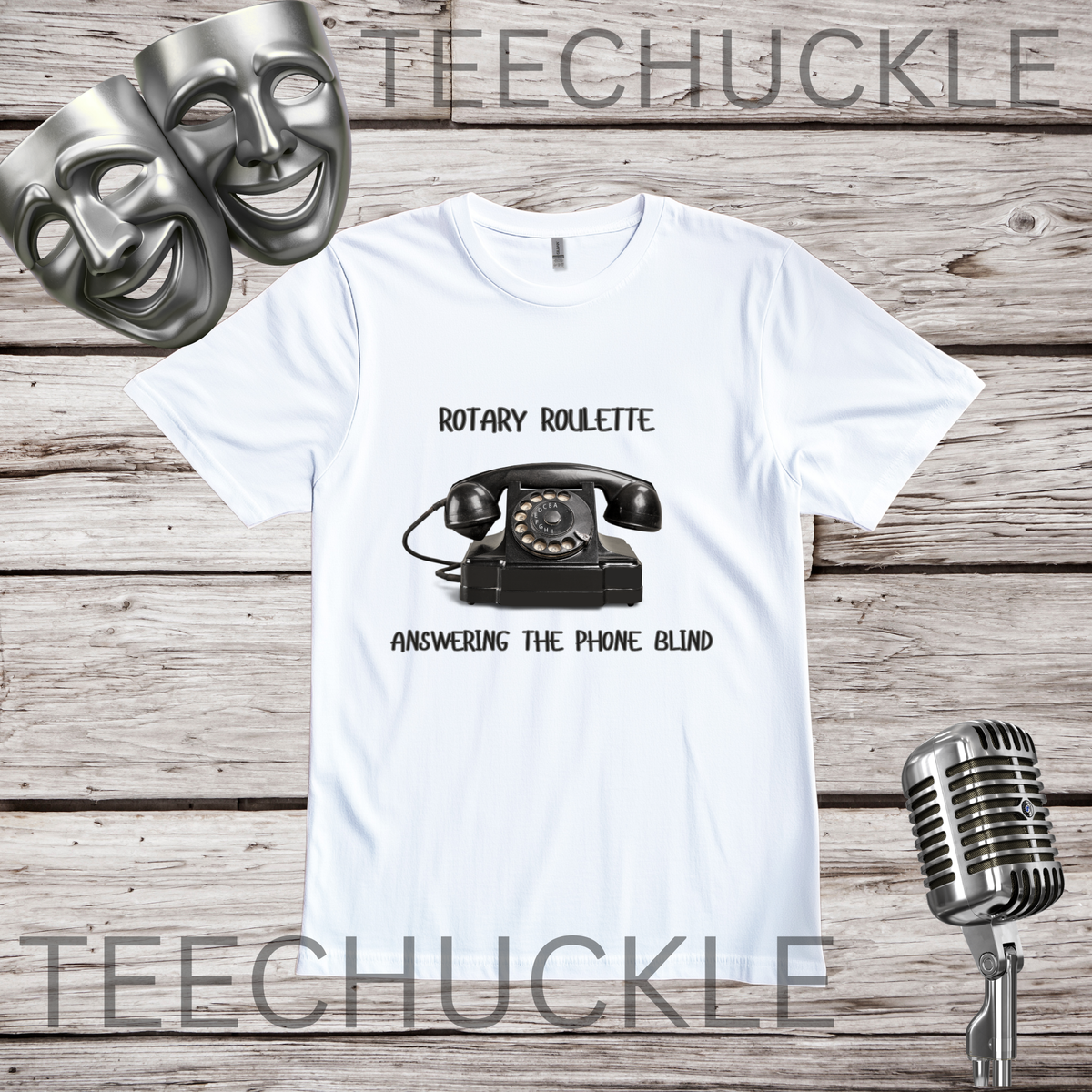A white graphic t-shirt featuring a black rotary telephone and the text 'ROTARY ROULETTE' and 'ANSWERING THE PHONE BLIND'. The t-shirt is displayed on a rustic wooden background with two silver comedy masks, a vintage microphone.