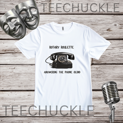 A white graphic t-shirt featuring a black rotary telephone and the text 'ROTARY ROULETTE' and 'ANSWERING THE PHONE BLIND'. The t-shirt is displayed on a rustic wooden background with two silver comedy masks, a vintage microphone.