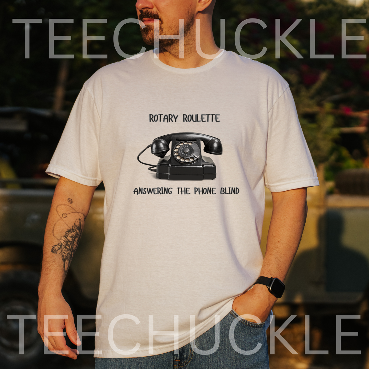A man wearing a white graphic t-shirt with a black rotary telephone print and the text 'ROTARY ROULETTE' and 'ANSWERING THE PHONE BLIND'. The background is a blurred outdoor setting