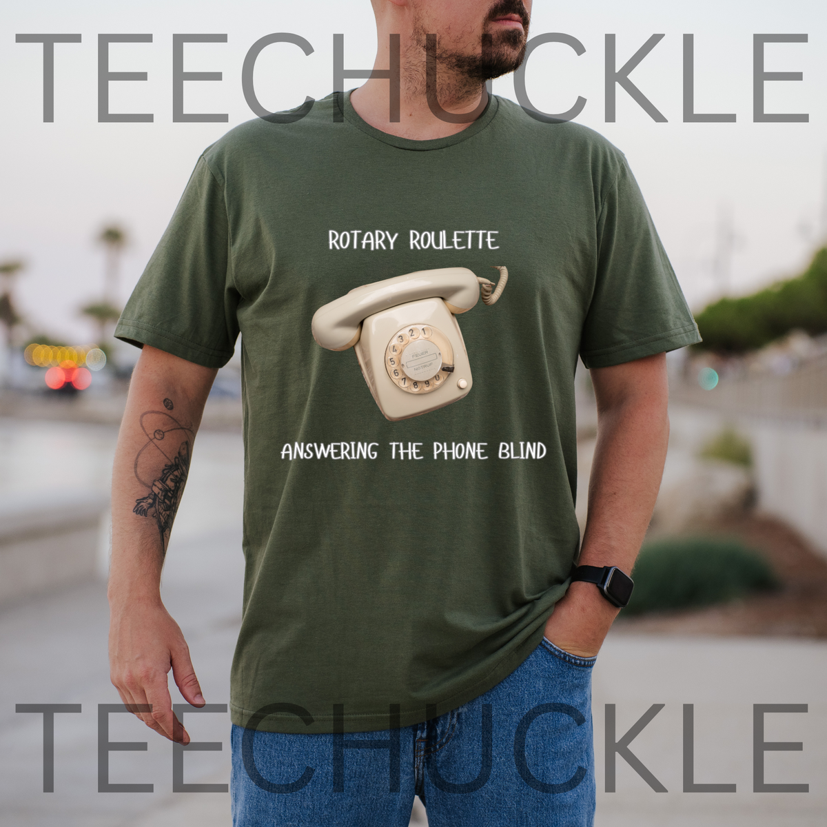 A man wearing a green graphic t-shirt with a retro rotary phone design and the text 'ROTARY ROULETTE ANSWERING THE PHONE BLIND'. The background is a blurred outdoor street scene.
