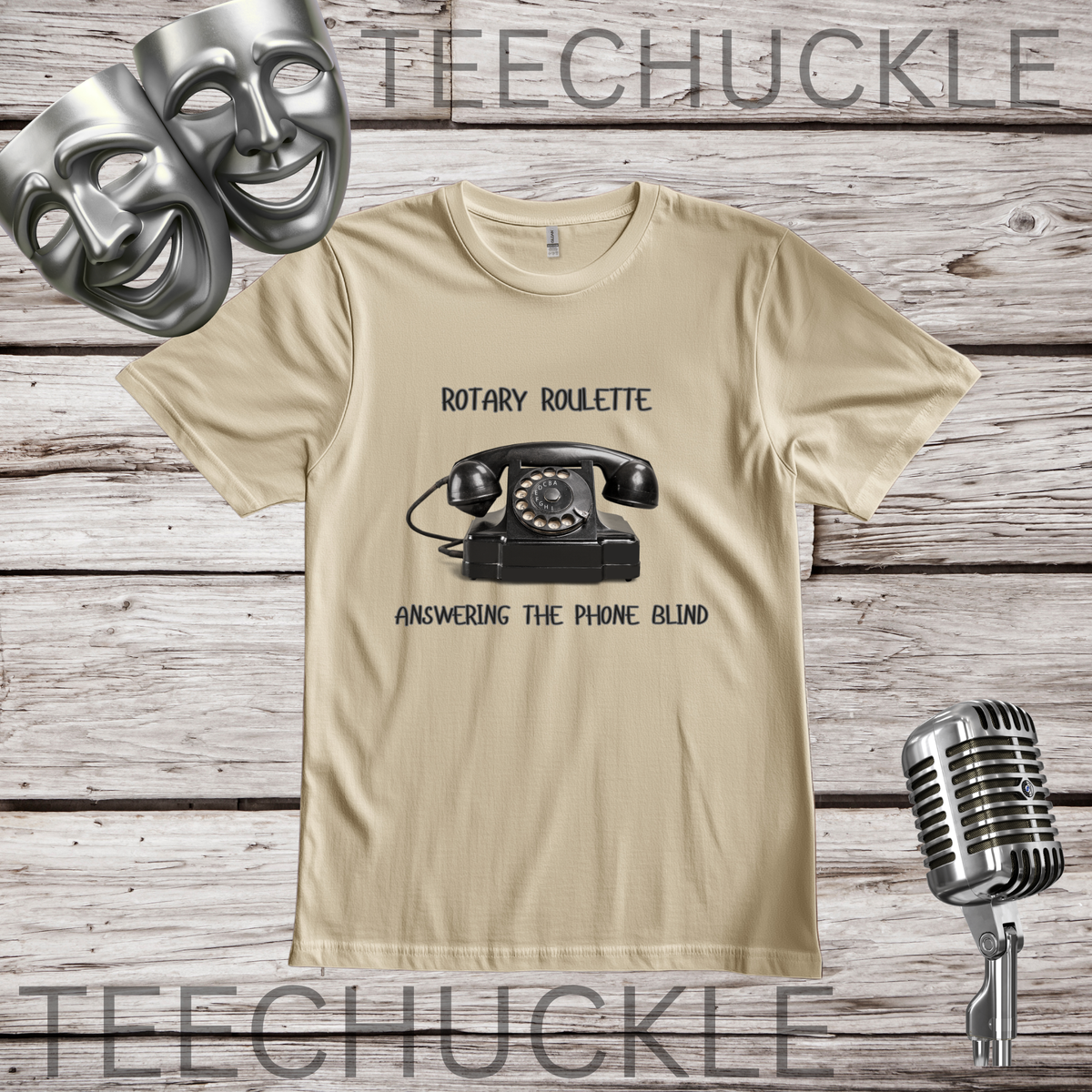 A sand graphic t-shirt featuring a black rotary telephone and the text 'ROTARY ROULETTE' and 'ANSWERING THE PHONE BLIND'. The t-shirt is displayed on a rustic wood background with silver comedy and microphone masks, a vintage microphone.