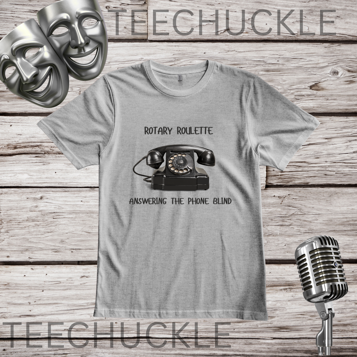 A  gray graphic t-shirt featuring a vintage rotary telephone and the text 'ROTARY ROULETTE' and 'ANSWERING THE PHONE BLIND'. The shirt is displayed on a rustic wooden background with silver theater masks, a vintage microphone.