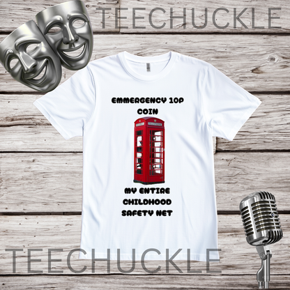 A white graphic t-shirt displayed on a rustic wood background, featuring a red telephone booth graphic and the text 'EMERGENCY 10P COIN MY ENTIRE CHILDHOOD SAFETY NET'. Two silver theatrical masks are in the top-left corner, and a vintage silver microphone is in the bottom-right.