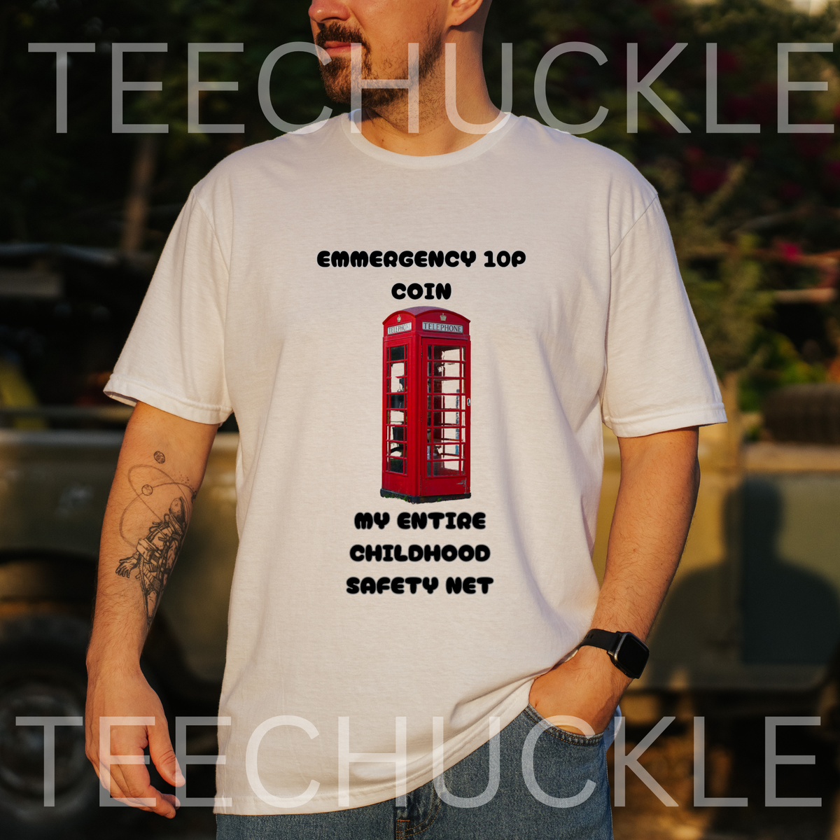 A man wearing a white graphic t-shirt with a red telephone booth print and black text reading 'EMERGENCY 100 COIN MY ENTIRE CHILDHOOD SAFETY NET'. The background is a blurred outdoor setting with warm sunset lighting