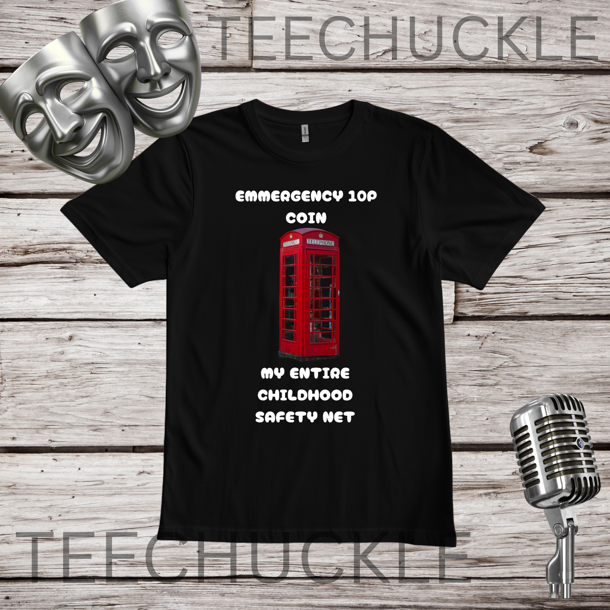 A black short-sleeve graphic T-shirt laid flat on a rustic wood plank background, with two silver theater masks, a vintage microphone. The shirt graphic shows a red telephone booth and white text reading 'EMERGENCY 109 COIN MY ENTIRE CHILDHOOD SAFETY NET'.