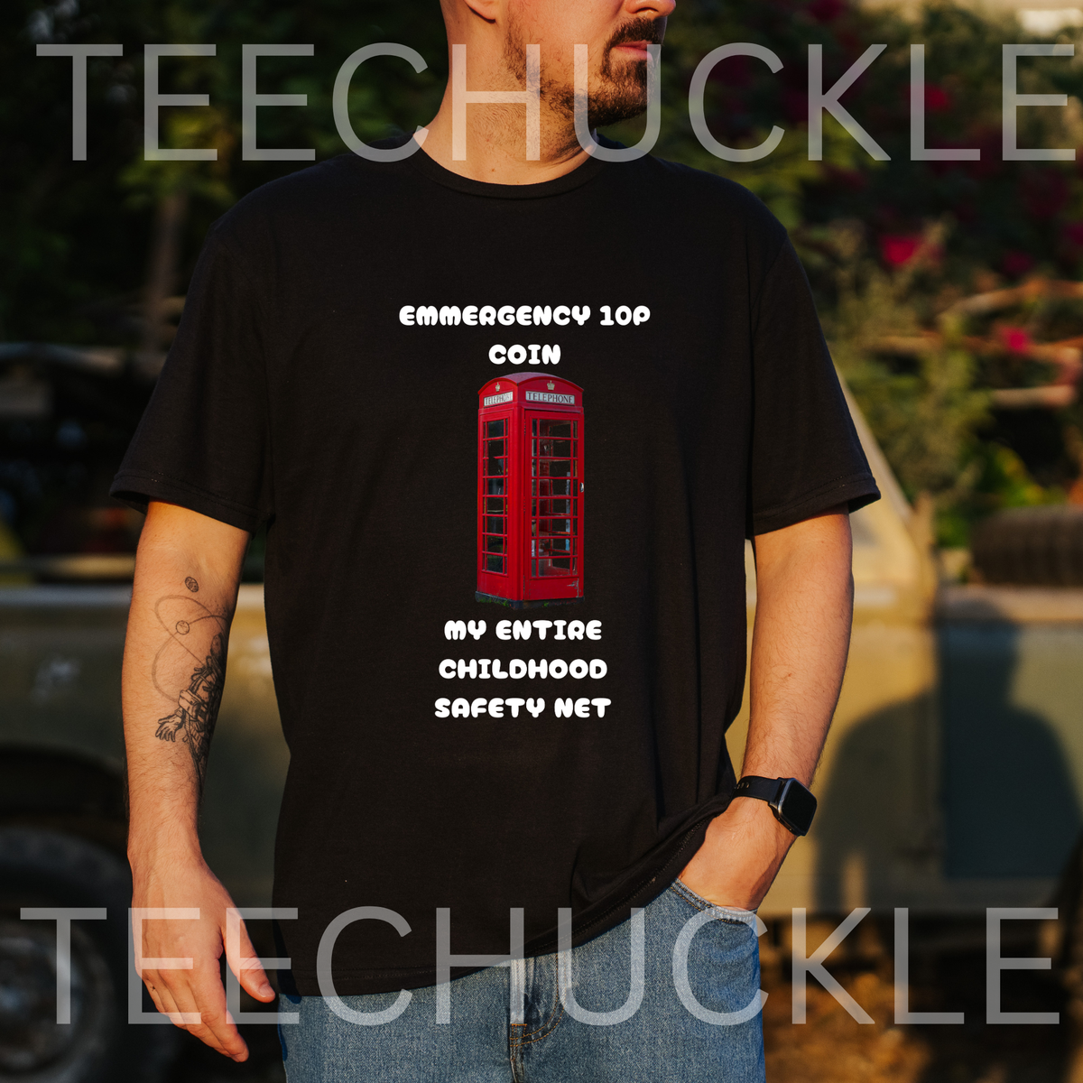 A man wearing a black graphic t-shirt with a red telephone booth and white text that reads 'EMERGENCY 10P COIN MY ENTIRE CHILDHOOD SAFETY NET'. The background is an outdoor blurred setting with a car and foliage