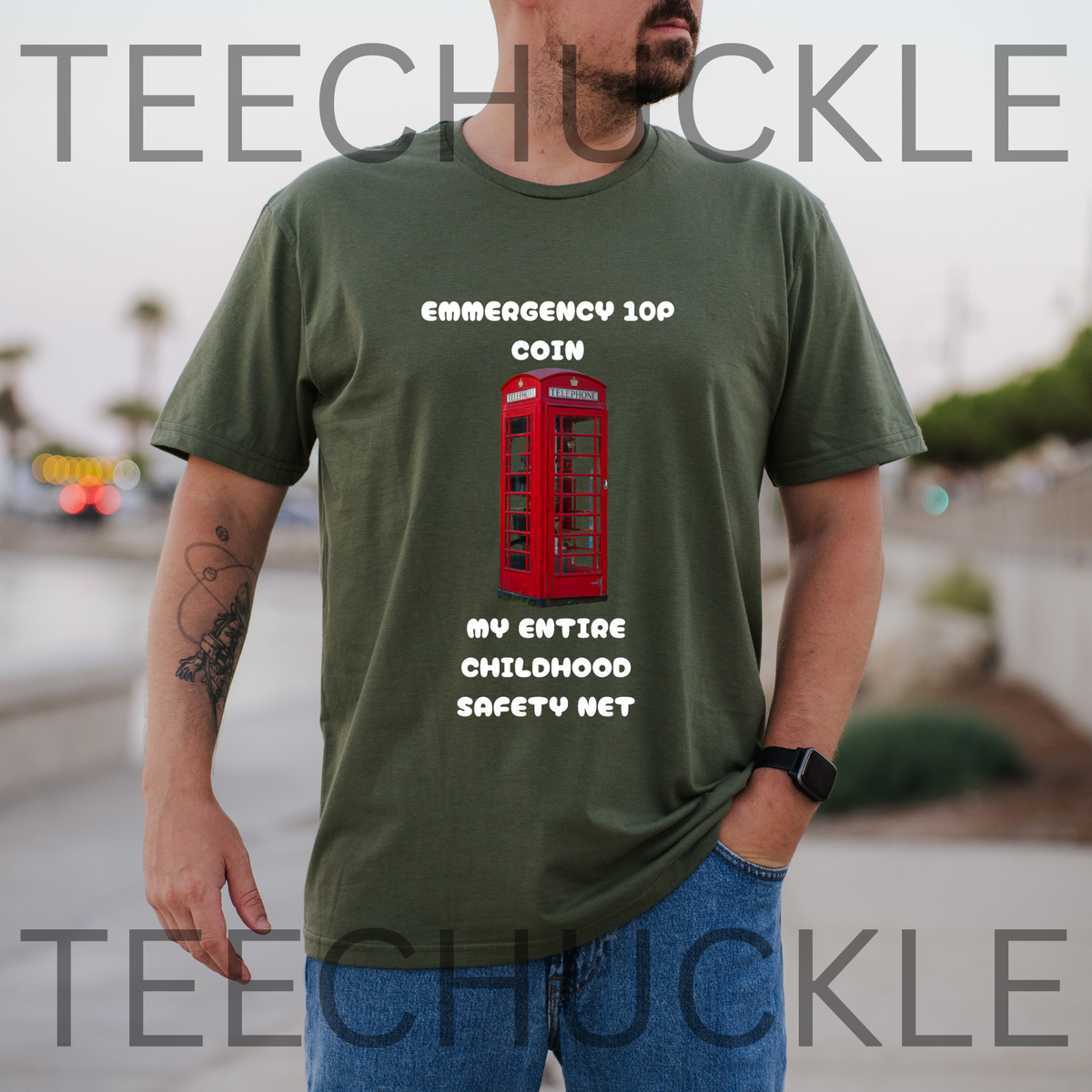 A man wearing a green graphic t-shirt with a red telephone booth and white text that reads 'EMERGENCY 10C COIN MY ENTIRE CHILDHOOD SAFETY NET'. The background is a blurred outdoor street scene.