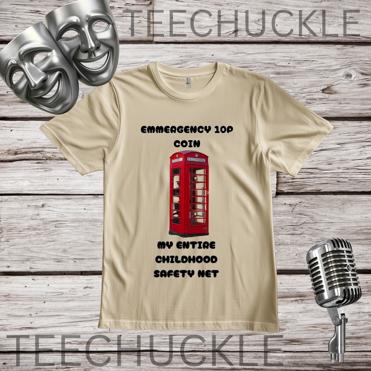 A SAND graphic t-shirt displayed on a rustic wood background, featuring a red telephone booth graphic and the text 'EMERGENCY 10P COIN MY ENTIRE CHILDHOOD SAFETY NET'. Two silver theatrical masks are in the top-left corner, and a vintage silver microphone is in the bottom-right.