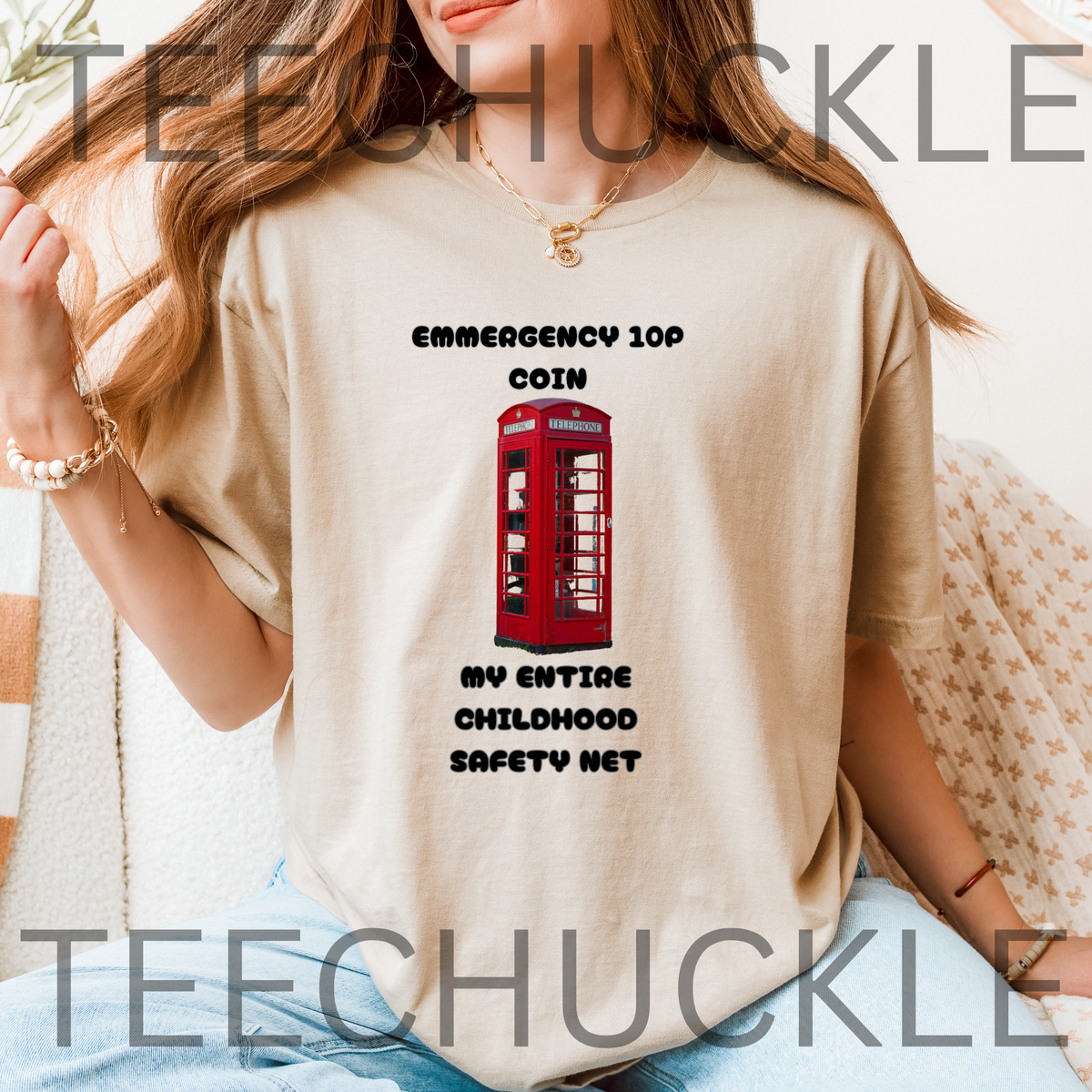 A woman wearing a sand graphic t-shirt with a red telephone booth and black text that reads 'EMERGENCY 10P COIN MY ENTIRE CHILDHOOD SAFETY NET'.