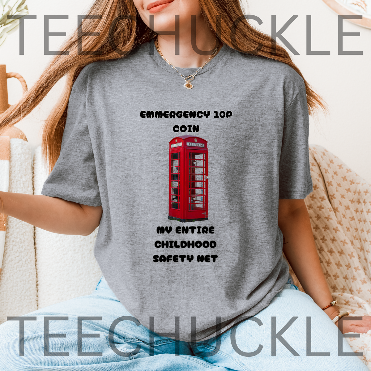 A woman sat wearing a grey graphic t-shirt displayed on a rustic wood background, featuring a red telephone booth graphic and the text 'EMERGENCY 10P COIN MY ENTIRE CHILDHOOD SAFETY NET'. 