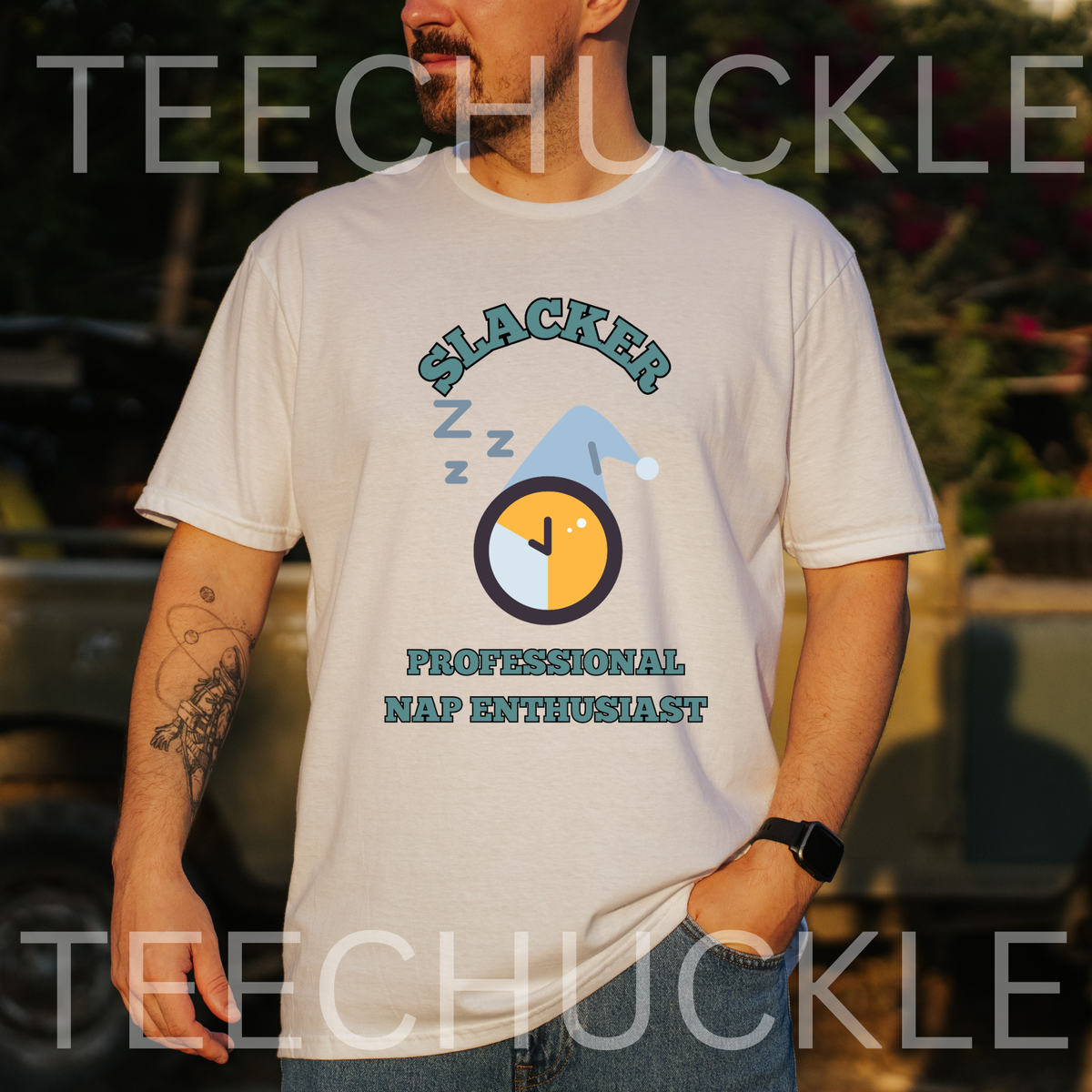 A man wearing a white graphic t-shirt with a 'Slacker Professional Nap Enthusiast' design, standing outdoors in front of a blurred vehicle background. The t-shirt features a cartoon sleeping clock and nap icon with the text 'SLACKER' and 'PROFESSIONAL NAP ENTHUSIAST'.