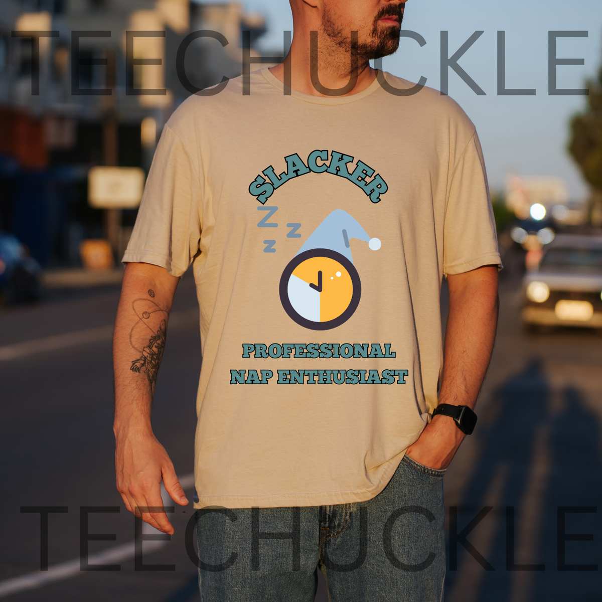 A man wearing a sand graphic t-shirt with a 'Slacker Professional Nap Enthusiast' design, standing outdoors on a street at sunset. The t-shirt features a cartoon clock with a blue sleep hat and 'Z' marks.