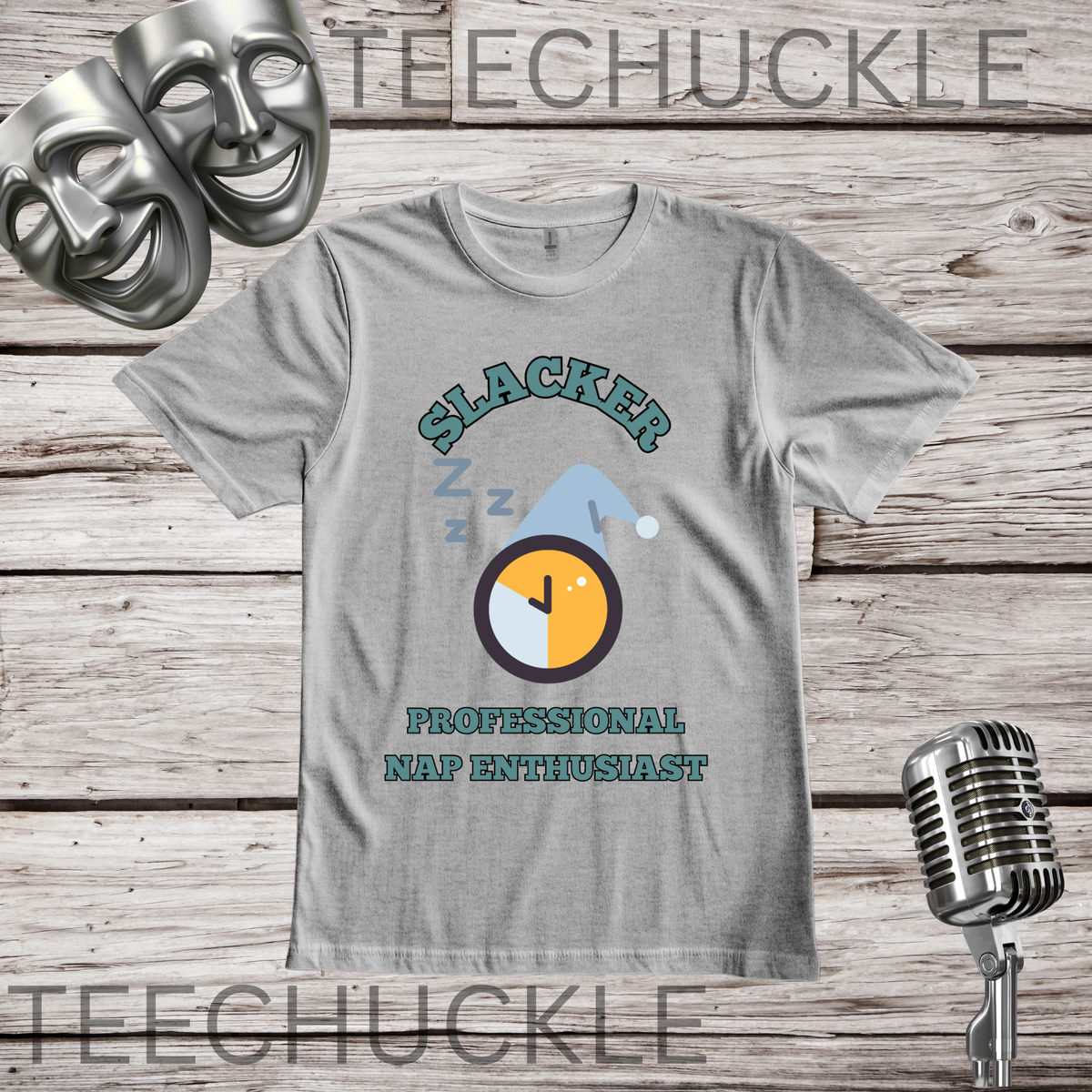 A gray graphic t-shirt displayed on a rustic wooden background, featuring a 'Slacker Professional Nap Enthusiast' design with a sleeping clock and blue pajama. The image includes decorative props like a vintage microphone, comedy masks.