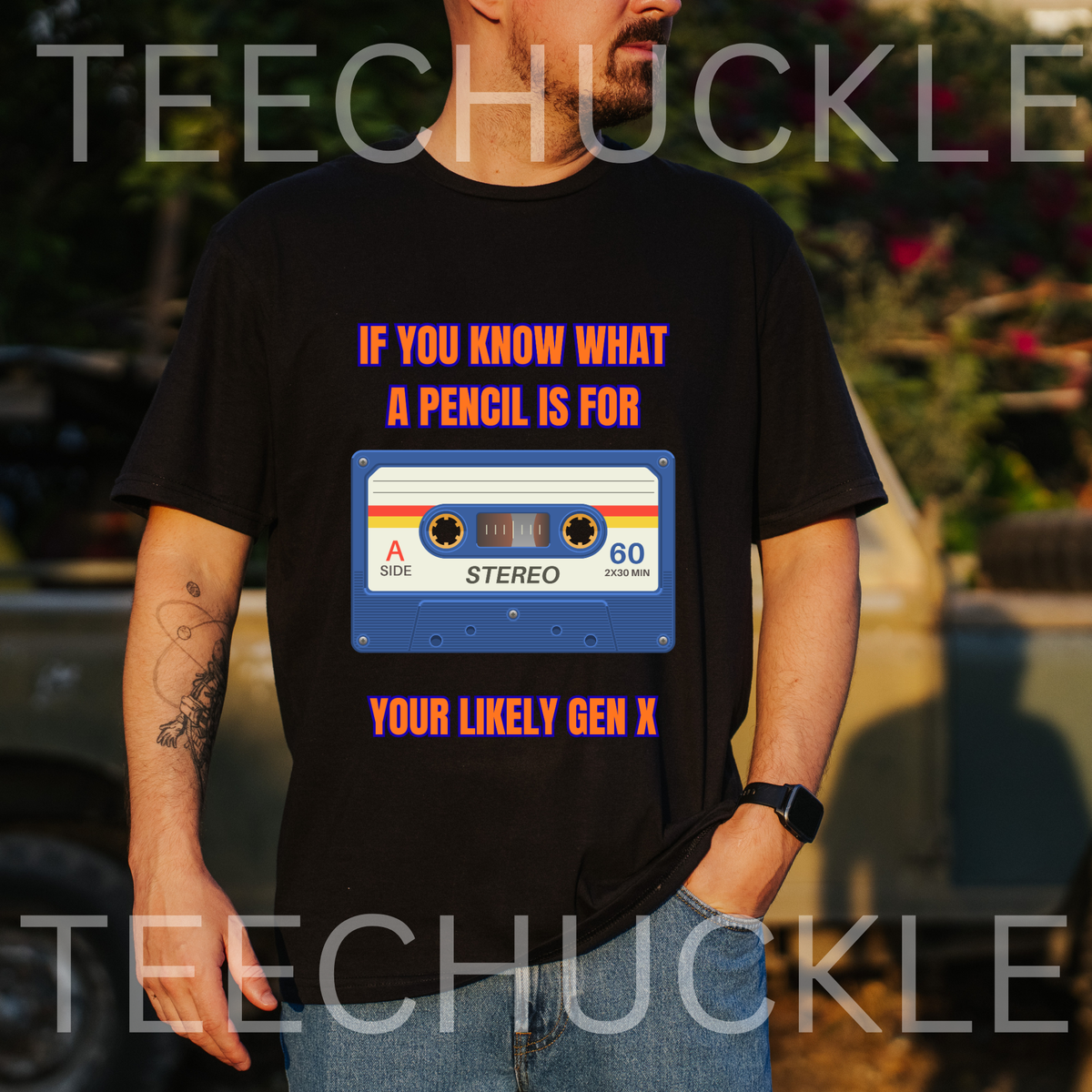 A man wearing a black graphic t-shirt with a retro cassette tape design and the text 'IF YOU KNOW WHAT A PENCIL IS FOR YOUR LIKELY GEN X'. The background is a blurred outdoor setting