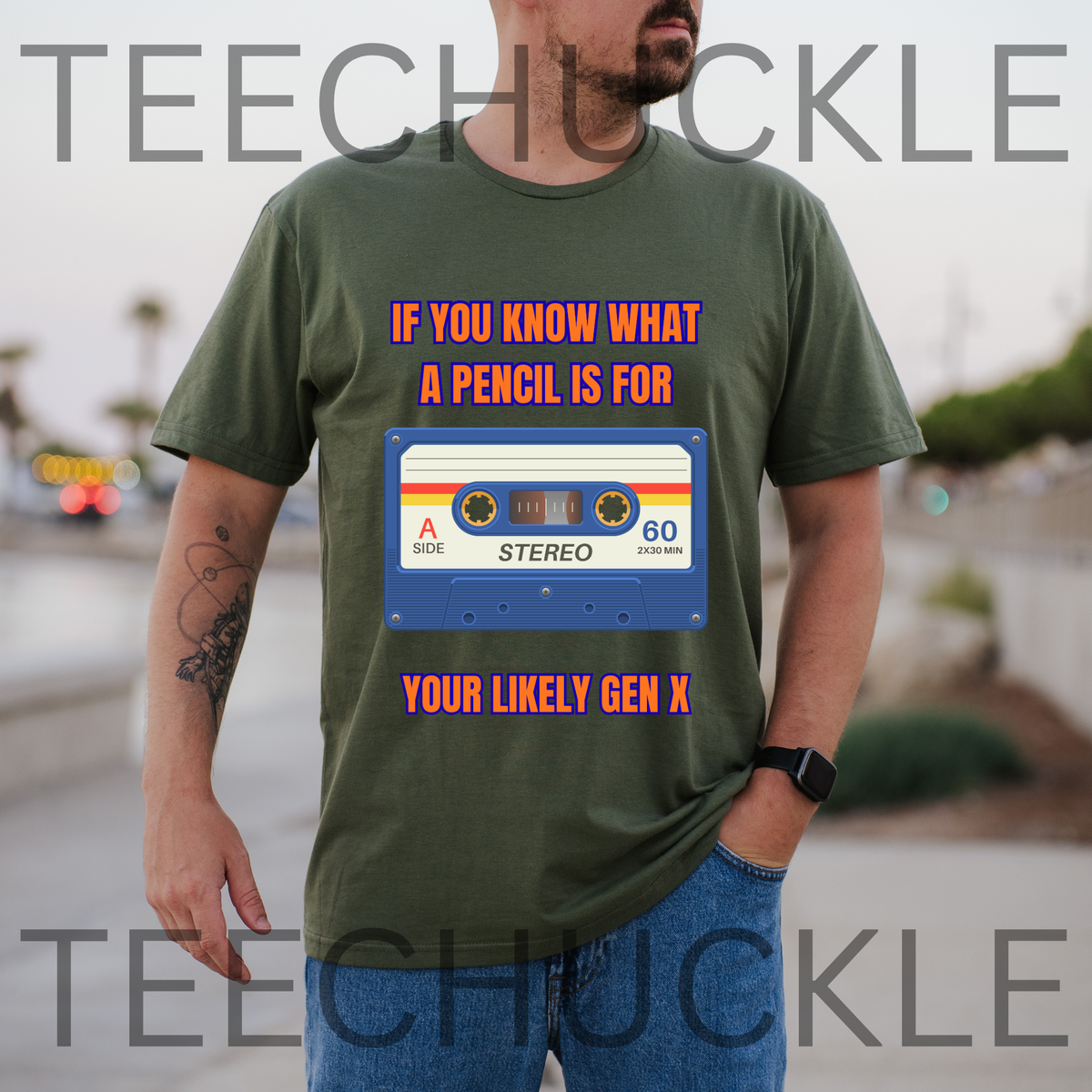 A man wearing a green graphic t-shirt with a retro cassette tape design and orange text that reads 'IF YOU KNOW WHAT A PENCIL IS FOR YOUR LIKELY GEN X'. The background is a blurred outdoor street scene.