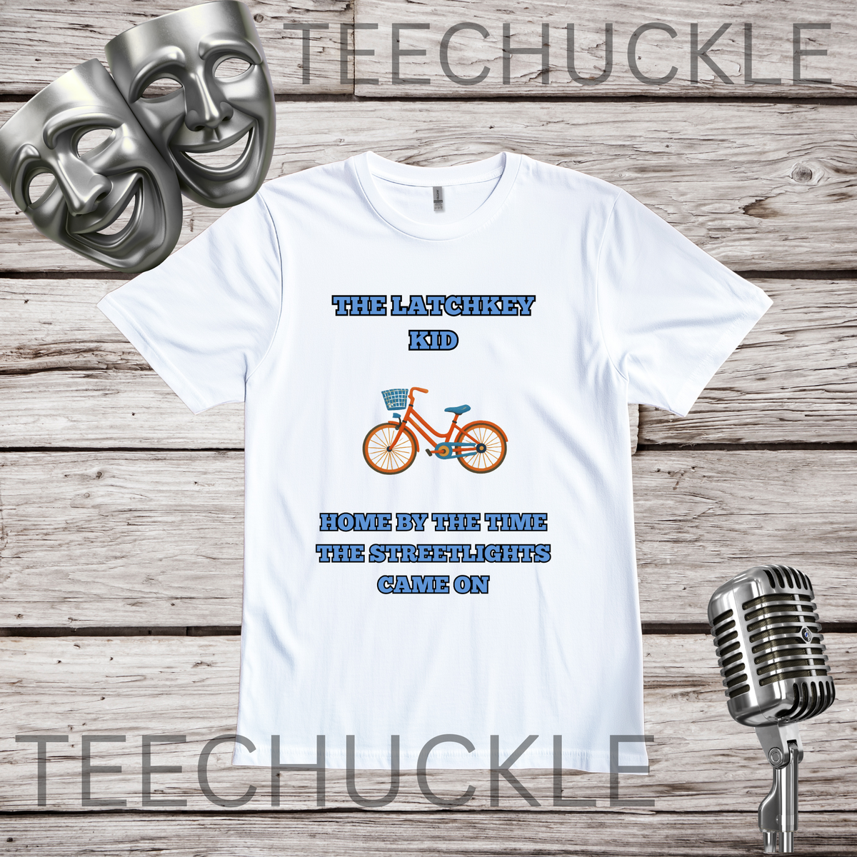 A white graphic t-shirt displayed on a rustic wood background, featuring the text 'THE LATCHKEY KID' and 'HOME BY THE TIME THE STREETLIGHTS CAME ON' in blue, with an orange bicycle graphic in the center. Two silver comedy masks and a vintage microphone are styled as props.