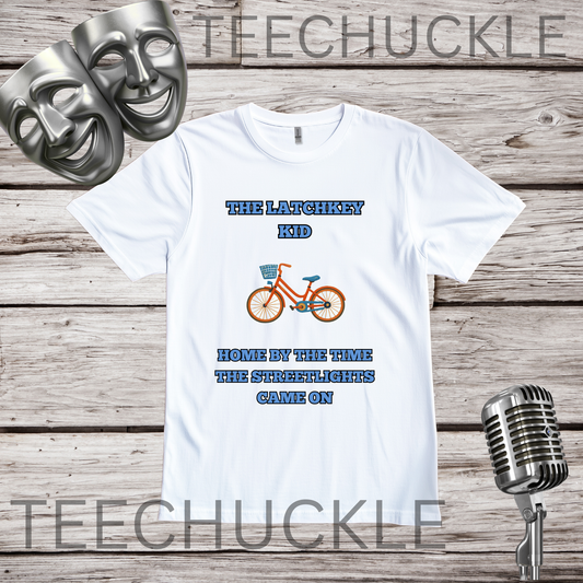 A white graphic t-shirt displayed on a rustic wood background, featuring the text 'THE LATCHKEY KID' and 'HOME BY THE TIME THE STREETLIGHTS CAME ON' in blue, with an orange bicycle graphic in the center. Two silver comedy masks and a vintage microphone are styled as props.