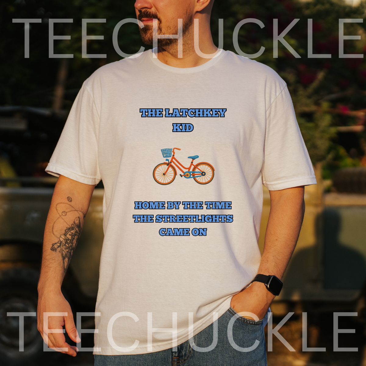 A man wearing a white graphic t-shirt with a blue and orange bicycle design and the text 'THE LATCHKEY KID HOME BY THE TIME THE STREETLIGHTS CAME ON', and is taken in an outdoor setting.