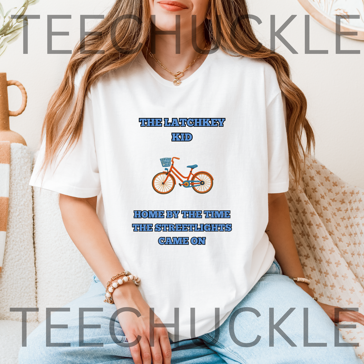 A woman wearing a white graphic t-shirt with blue and orange text and a bicycle illustration, sitting in a cozy indoor setting. The t-shirt features the text 'THE LATCHKEY KID HOME BY THE TIME THE STREETLIGHTS GAME ON' around a retro orange bicycle graphic.