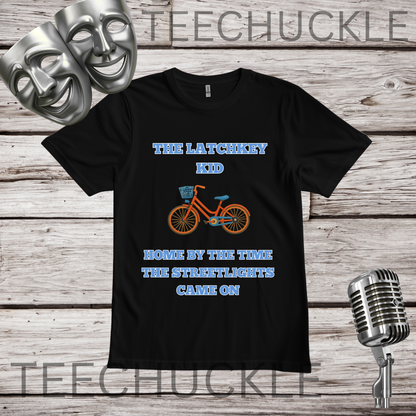 A black short-sleeve graphic T-shirt laid flat on a rustic wood plank background, with theatrical comedy and tragedy masks, a vintage microphone. The shirt has blue text reading 'THE LATCHKEY KID' and 'HOME BY THE TIME THE STREETLIGHTS CAME ON' with an orange bicycle graphic in the center.