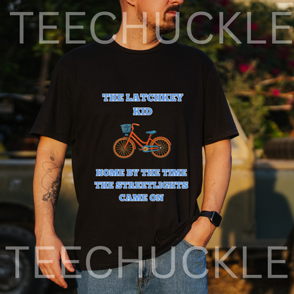 A man wearing a black graphic t-shirt with a blue and orange bicycle design and the text 'THE LATCHKEY KID HOME BY THE TIME THE STREETLIGHTS CAME ON', and the background is a blurred outdoor setting.