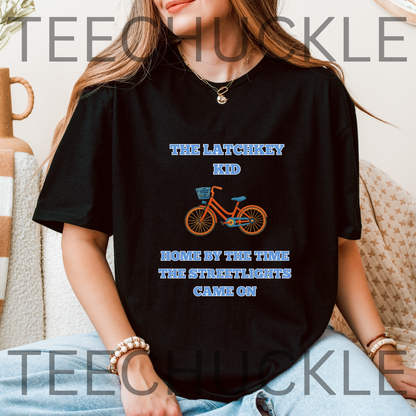 A woman wearing a black graphic t-shirt with a blue and orange bicycle design and the text 'THE LATCHKEY KID HOME BY THE TIME THE STREETLIGHTS GAVE ON!'. 