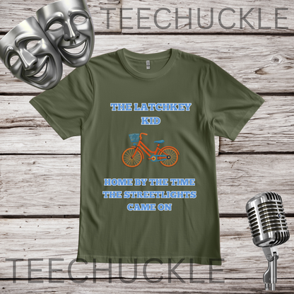 A green graphic t-shirt displayed on a rustic wood background, flanked by silver theater masks on the top left and a vintage microphone on the bottom right. The t-shirt features a graphic of an orange bicycle with blue text that reads 'THE LATCHKEY KID HOME BY THE TIME THE STREETFLIGHTS CAME ON'.