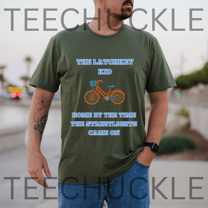 A man wearing a green graphic t-shirt with a blue and orange bicycle design and the text 'THE LATCHKEY KID HOME BY THE TIME THE STREETLIGHTS CAME ON', and is set against a blurred outdoor background.