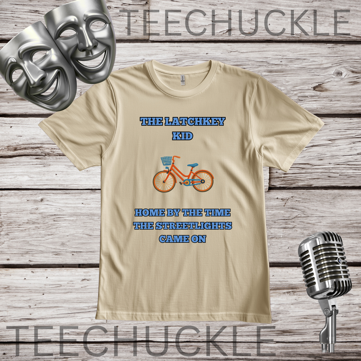 A sand graphic t-shirt displayed on a rustic wood background, featuring a vintage bicycle illustration and the text 'THE LATCHKEY KID HOME BY THE TIME THE STREETLIGHTS CAME ON'. The shirt is styled with silver comedy and tragedy masks on the top left, a vintage microphone on the bottom right.