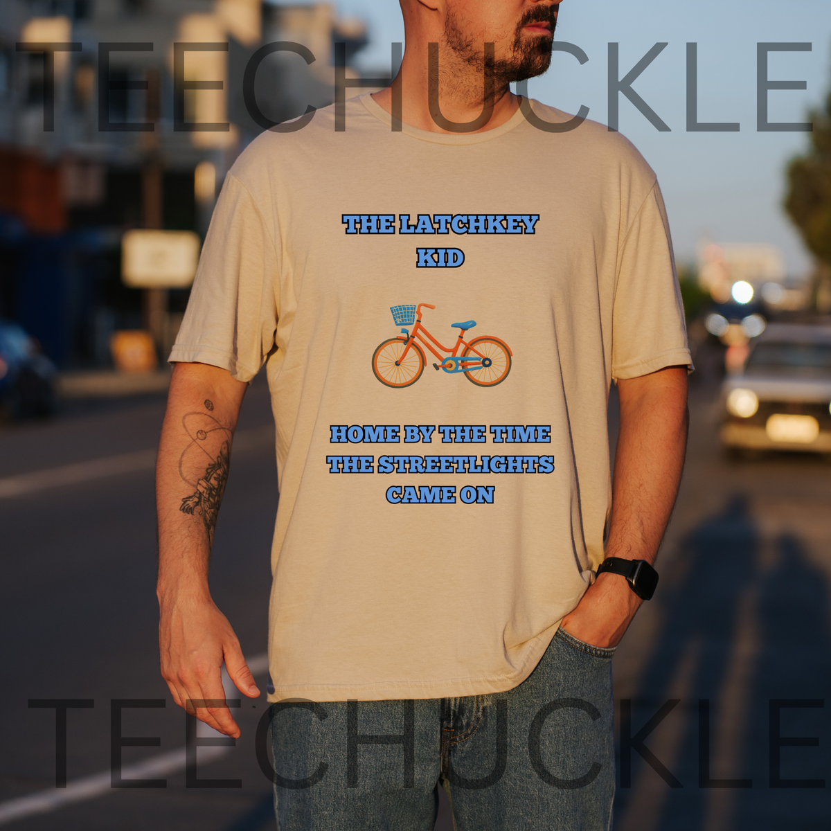 A man wearing a sand graphic t-shirt with blue and orange bicycle illustration, standing outdoors on a street at sunset. The shirt features the text 'THE LATCHKEY KID HOME BY THE TIME THE STREETLIGHTS CAME ON'.