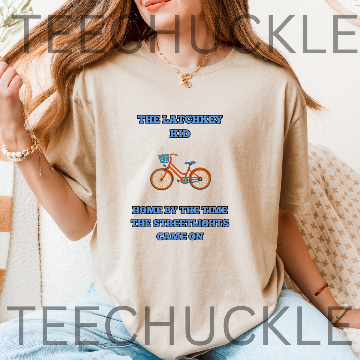 A woman wearing a sand graphic t-shirt with a blue and orange bicycle design and the text 'THE LATCHKEY KID HOME BY THE TIME THE STREETLIGHTS GAME ON'.