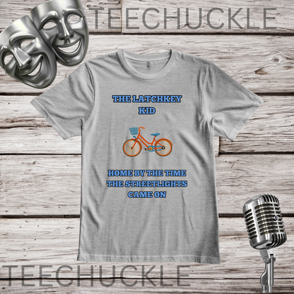 A gray graphic t-shirt displayed on a rustic wood background with props including two silver theater masks, a vintage microphone. The t-shirt features a graphic of an orange bicycle with blue text that reads 'THE LATCHKEY KID HOME BY THE TIME THE STREETLIGHTS GAME ON'.