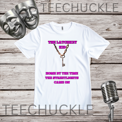 A white short-sleeve graphic T-shirt laid flat on a rustic wood plank background, with two silver theater masks in the top left and a vintage microphone in the bottom right. The shirt has a central graphic of a gold chain and key with pink text reading 'THE LATCHKEY KEY' and 'HOME BY THE TIME THE STREETLIGHTS CAME ON.